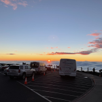 Visitors and parked vehicles at the Haleakala Visitor Center parking lot watch the sun setting above a cloud layer. Photograph credited to NPS / LM.