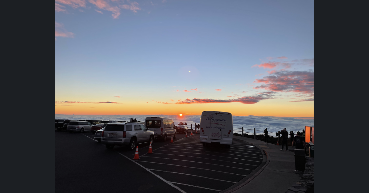 Visitors and parked vehicles at the Haleakala Visitor Center parking lot watch the sun setting above a cloud layer. Photograph credited to NPS / LM.
