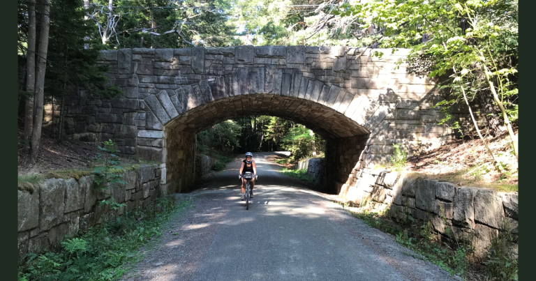 A helmeted bicyclist rides beneath a stone carriage road bridge surrounded by trees in Acadia National Park